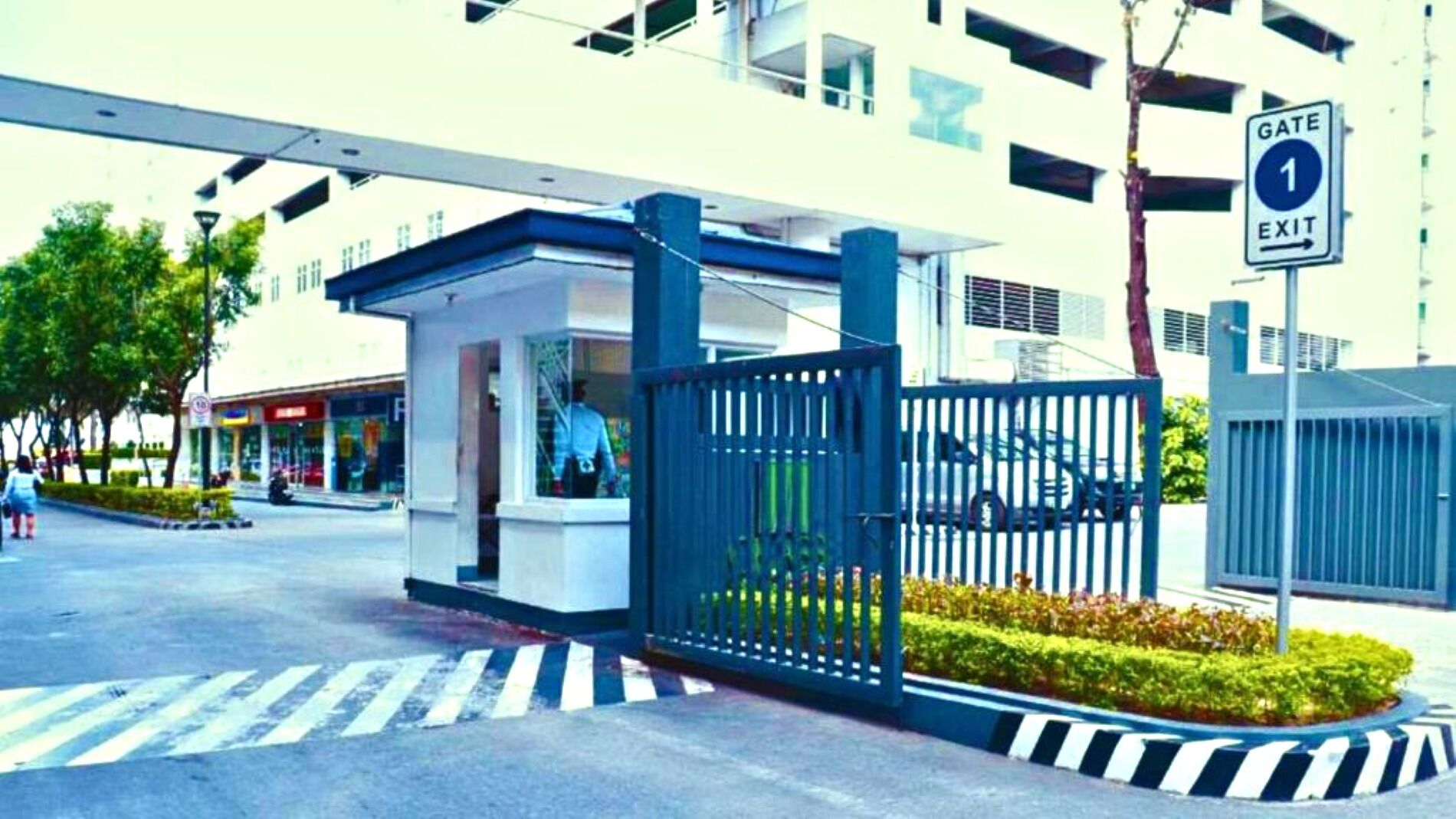 A gated entrance with a blue fence, a pedestrian crossing, and a building with multiple windows in the background.