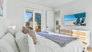Et soveværelse med en stor seng, en loftsvifte, et fjernsyn der viser et strandbillede og udsigt til det fri gennem en skydedør.