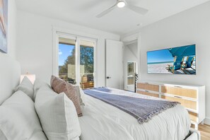 Ein Schlafzimmer mit einem großen Bett, einem Deckenventilator, einem Fernseher, der eine Strandansicht zeigt, und Blick ins Freie durch eine Schiebetür.