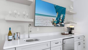 Et moderne køkken med et stort fladskærms-tv, der viser et strandmotiv, en vinflaske og en kaffemaskine.