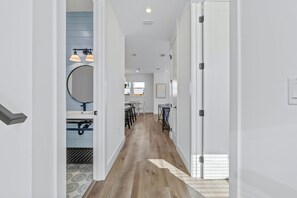 Un corridoio con pavimento in legno, pareti bianche e una porta che conduce a una stanza con uno specchio rotondo e lampade a sospensione.