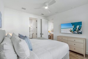 Quarto com cama, ventoinha no teto, cômoda e televisão na parede exibindo uma cena de praia.