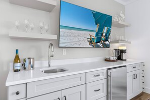 A modern kitchen with a large flat-screen TV displaying a beach scene, a wine bottle, and a coffee maker. - Blue Pointe: Private heated pool, Free golf cart, 1min 2 beach, Game & Bunk room (Miramar Beach)