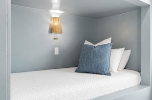 A single bed with a white quilted mattress, a blue pillow, and a white pillow. There is a wall-mounted lamp with a woven shade. - Gulf front | Heated Private Pool |Sleeps 18 | Bonfire Incl (Panama City Beach)