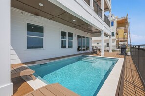 Un área de piscina con piscina azul cristalina, terraza de madera y un edificio blanco con amplias ventanas.