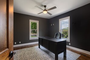 Ein Zimmer mit Schreibtisch, zwei Fenstern, einem Deckenventilator und einem gemusterten Teppich.