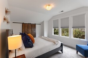Ein Schlafzimmer mit einem Bett, einem Nachttisch, einer Lampe, einem Fenster mit Jalousien und Blick auf Grünfläche außerhalb.