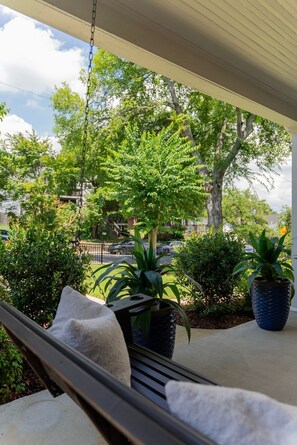 A porch with a swing chair, potted plants, and a view of a street with cars and trees. - 🌸 Magnolia House @ 12 South | Private Pool | Sleeps 10 | Fireplace (Nashville)