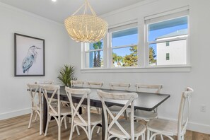 Ein Esszimmer mit einem großen Tisch und sechs Stühlen, einer Pendelleuchte, einem gerahmten Kunstwerk sowie Fenstern mit Blick auf ein Gebäude und Bäume.