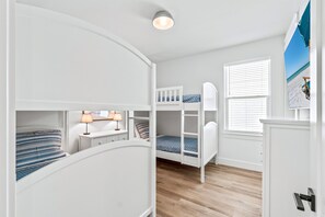 Ein Kinderzimmer mit Etagenbett, Schreibtisch und Lampe, einem Fenster mit Jalousien und einer Wanddekoration im Strandhausstil.