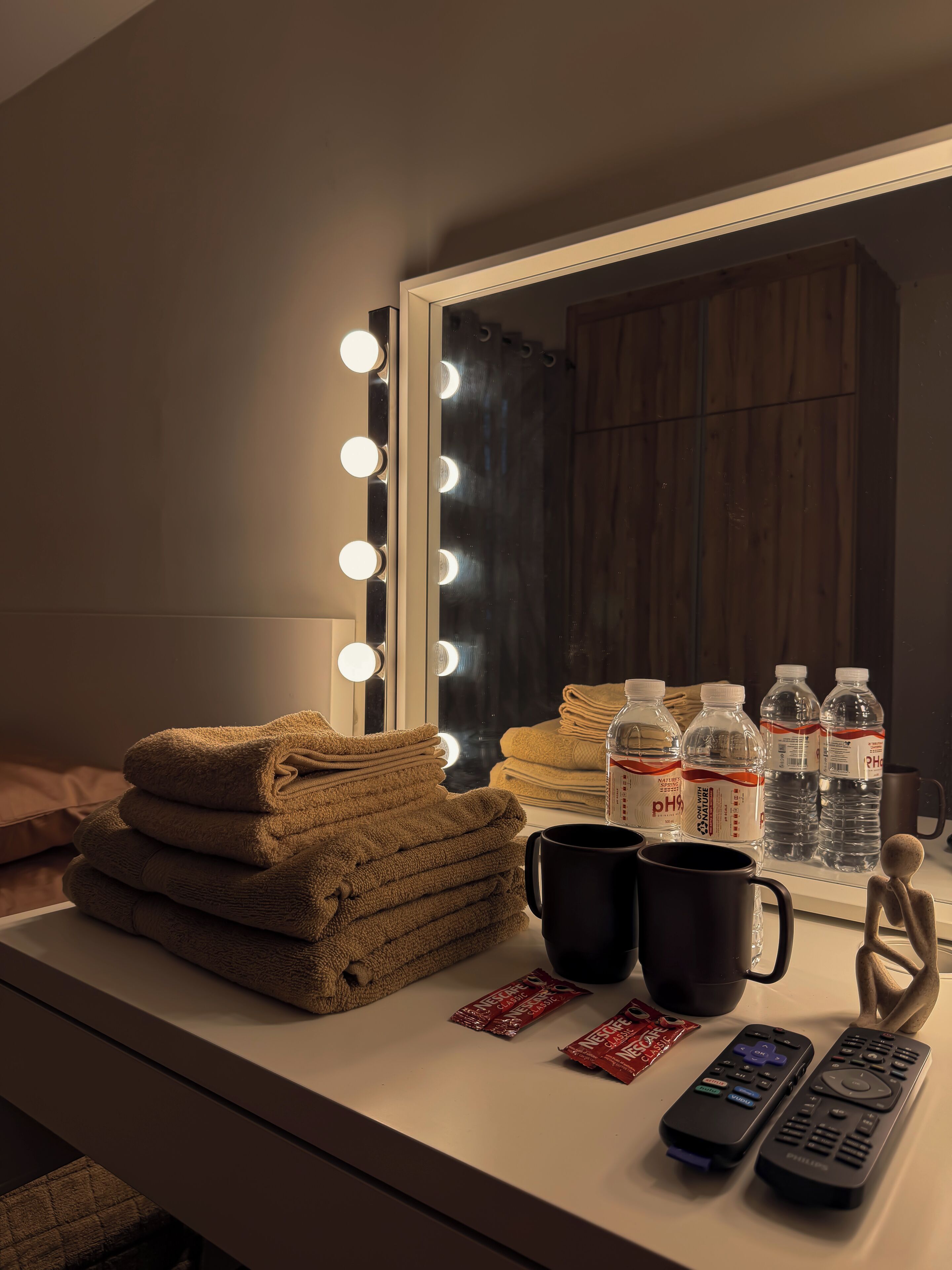 A vanity with a mirror, lights, towels, water bottles, a mug, and remote controls.