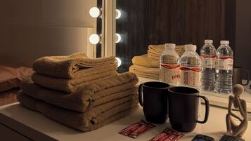 A vanity with a mirror, lights, towels, water bottles, a mug, and remote controls.
