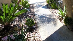 A pathway leading to a building with a textured exterior, surrounded by various plants and a tennis court in the background. - 2-bedroom condo-Fun Colorful-Fabulous Rancho Mirage- AC, pools, hot tubs, tennis (Rancho Mirage)