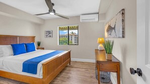 A bedroom with a bed, ceiling fan, window, artwork, and a desk with a lamp. - Modern Paradise | Kihei 2BR Near Beach (Kihei)