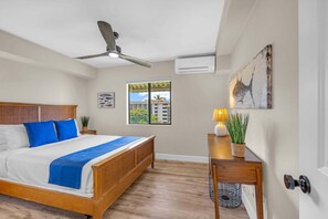 Ein Schlafzimmer mit Bett, Deckenventilator, Fenster, Kunstwerk und einem Schreibtisch mit Lampe.