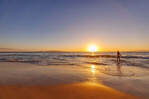 Een strandscène met iemand die in het water staat, de zon zakt in de verte.