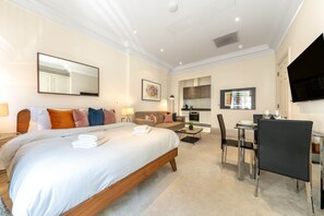 A hotel room with a large bed, a sofa, a dining table with chairs, a TV, and a kitchen area. - Royal Studio Apartment in Mayfair (London)