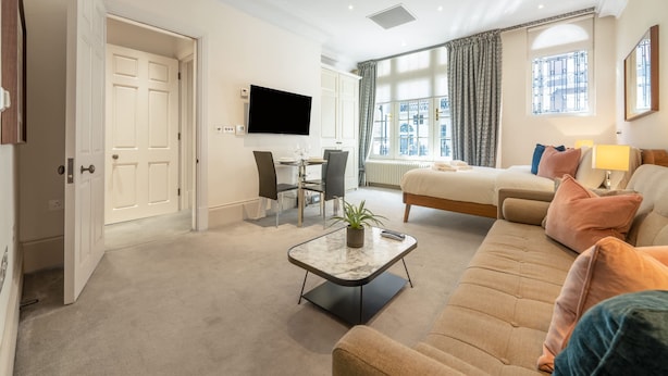 A modern hotel room with a sofa, a dining area with a table and chairs, a flat-screen TV mounted on the wall, and a large window with curtains. - Royal Studio Apartment in Mayfair (London)