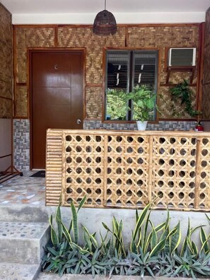 A wooden door, a window with blinds, a wall with a woven pattern, and a plant in a planter.