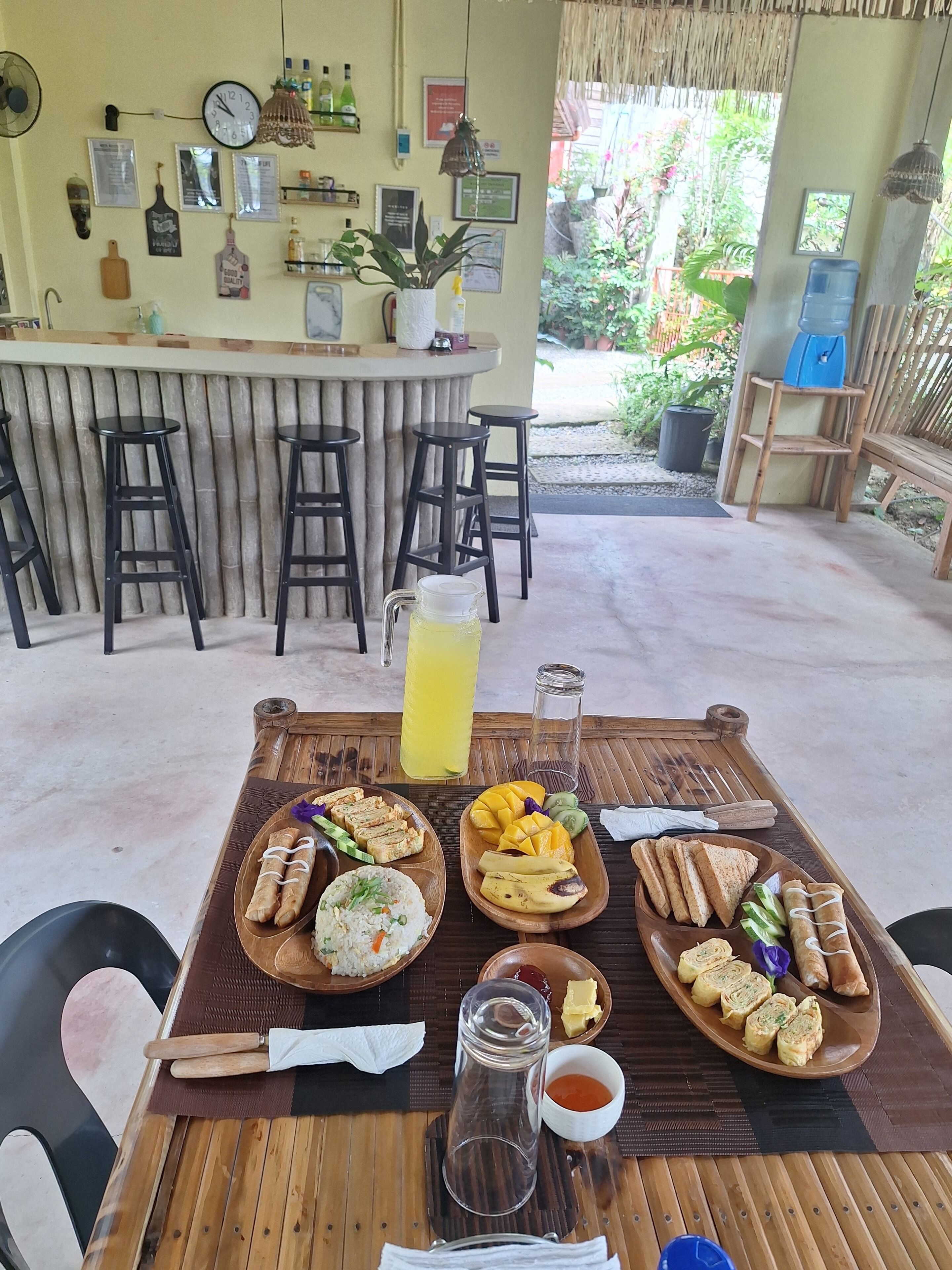 Una mesa de madera con diversos alimentos y una jarra de bebida amarilla, ubicada en un espacio con barra y taburetes.