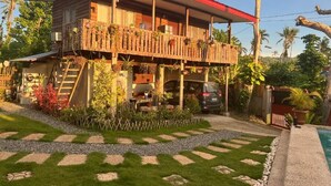 Une maison en bois à deux étages, avec un toit rouge, un balcon orné de plantes suspendues et une voiture garée dans l’allée.