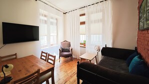 Ein Zimmer mit einem Holztisch, Stühlen, einer Couch, einem an der Wand befestigten Fernseher und einem Fenster mit Vorhängen.