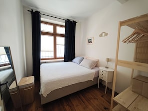 Un dormitorio con cama, una mesita de noche, lámpara, armario de madera, ventana con cortinas y un espejo.