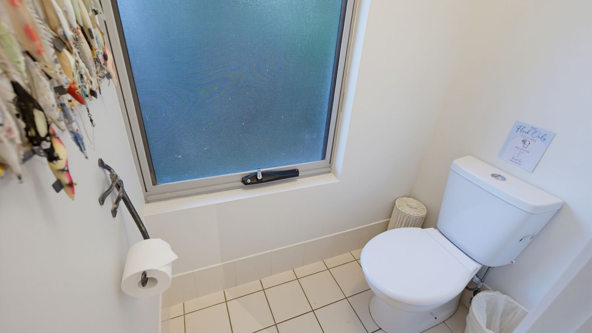 Une salle de bain comprenant des toilettes, une fenêtre avec des stores, un rouleau de papier toilette et une poubelle.