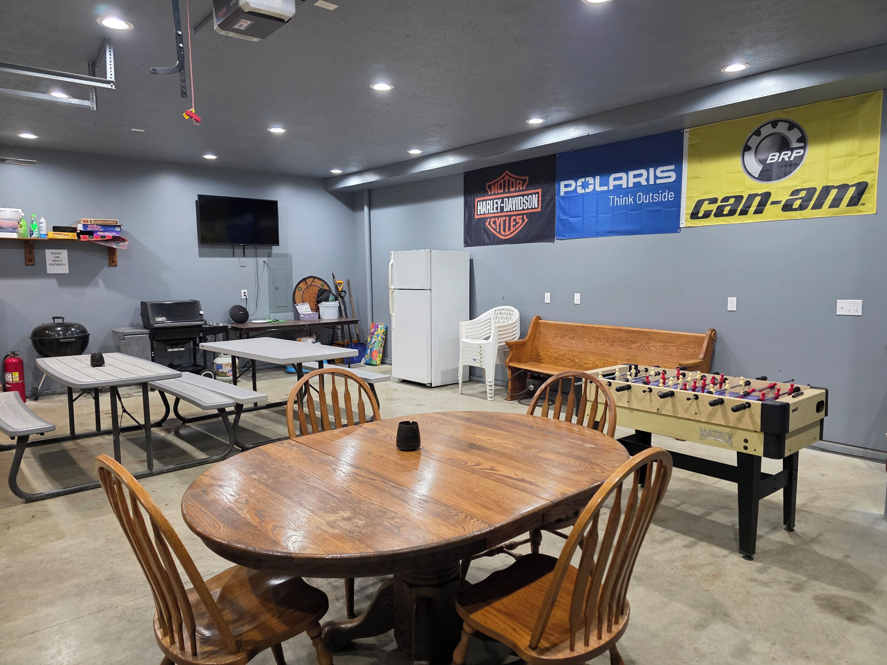 Une pièce comprenant une table ronde en bois, quatre chaises, un baby-foot, un barbecue, un réfrigérateur et des bannières aux couleurs de Harley-Davidson, Polaris et Can-Am.