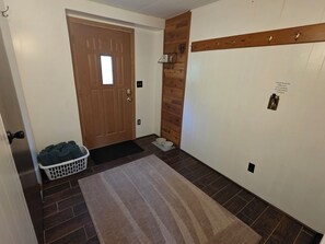 Un pasillo con una puerta de madera, una cesta de ropa, un tapete y un estante en la pared con un objeto pequeño.