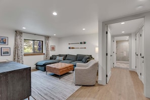 Ein modernes Wohnzimmer mit einer Couch, einem Hocker und einem Sessel, einem Holztisch und einem Teppich. Zwei Fenster mit Vorhängen, ein an der Wand befestigtes Stand-Up-Paddlebrett und ein Flur, der zu einem Badezimmer führt.