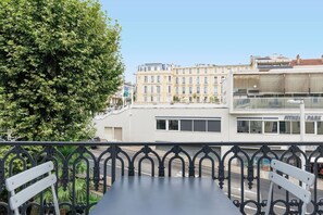 Ein Balkon mit Tisch und Stühlen, der auf eine Straße mit Gebäuden und einem Baum blickt.