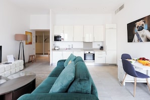 A modern living room with a teal sofa, a dining table with chairs, a kitchen area with white cabinets, and a painting on the wall. - Upstays Spacious 77m² T3 Apartment with Terrace (Cannes)
