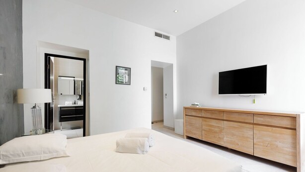 Ein modernes Hotelzimmer mit einem großen Bett, einer Holzkommode, einem Flachbildfernseher und einem durchgehenden Türspalt sichtbaren Badezimmer.