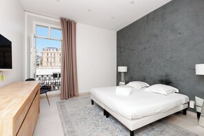 Ein modernes Schlafzimmer mit einem großen Bett, einem Holztisch, einem Balkon mit Blick auf Gebäude und einer grauen, strukturierten Wand.