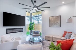 A modern living room with a flat-screen TV, a ceiling fan, a glass coffee table, a beige sofa, and a large window. - Downtown Escape | Modern Stay with Pool (Austin)
