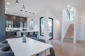 A modern kitchen with dark cabinets, a white countertop, and a dining area with a white table and grey chairs. - Downtown Escape | Modern Stay with Pool (Austin)
