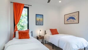 A bedroom with two beds, a ceiling fan, a window with orange curtains, a framed picture, and a nightstand with a lamp. - Downtown Escape | Modern Stay with Pool (Austin)