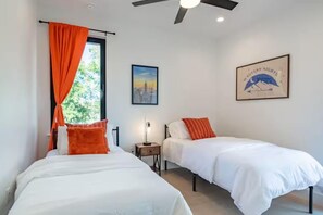 Ein Schlafzimmer mit zwei Betten, einem Deckenventilator, einem Fenster mit orangefarbenen Vorhängen, einem gerahmten Bild und einem Nachttisch mit Lampe.