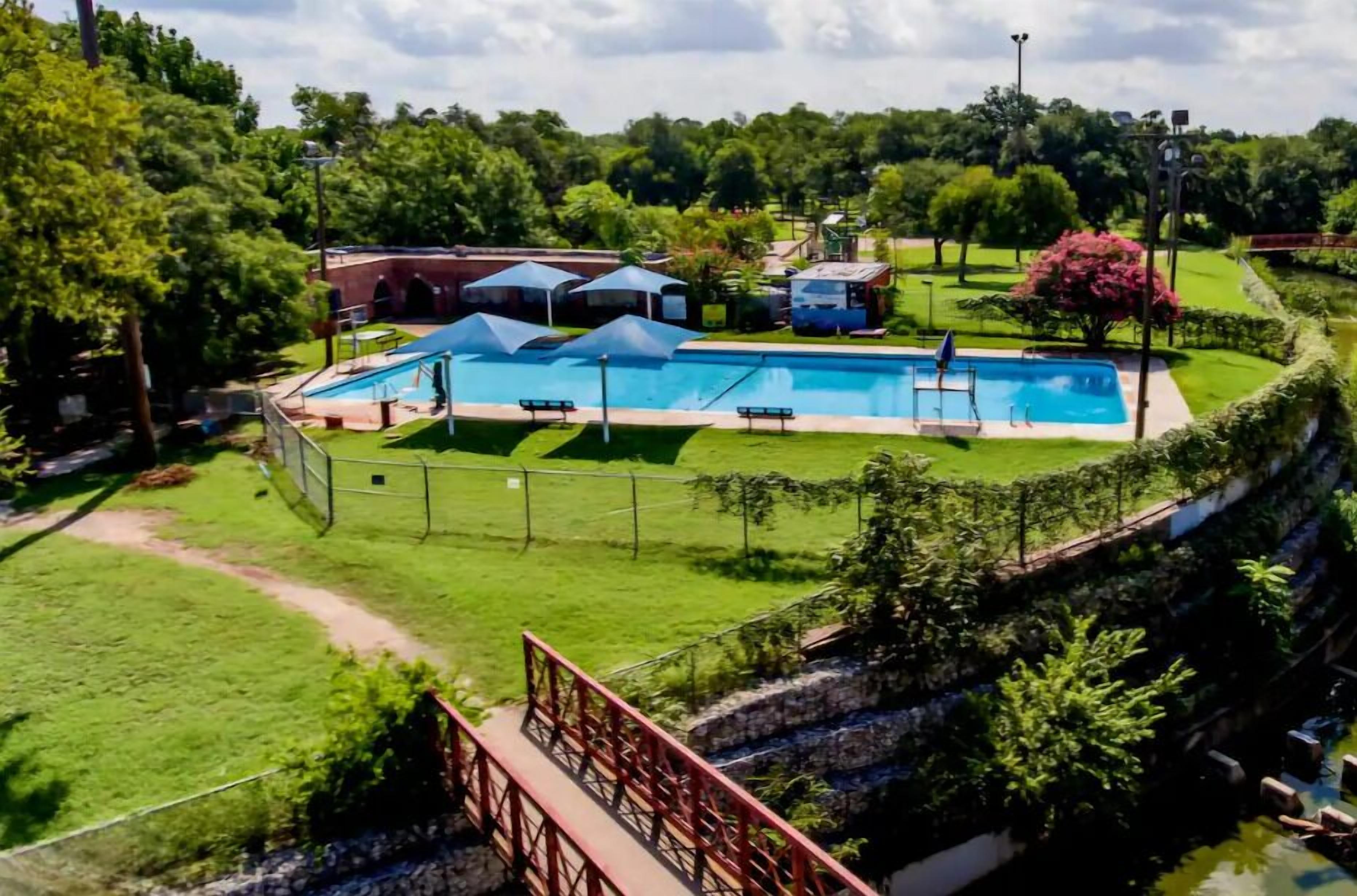 A swimming pool surrounded by a fenced area with a grassy lawn, benches, and a small building.