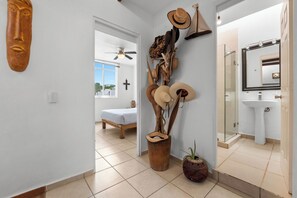 A hallway with a bed, a ceiling fan, a cross on the wall, and a bathroom with a sink and mirror. - Bucerias | 3 Bedrooms and Private Patio (Bucerías)