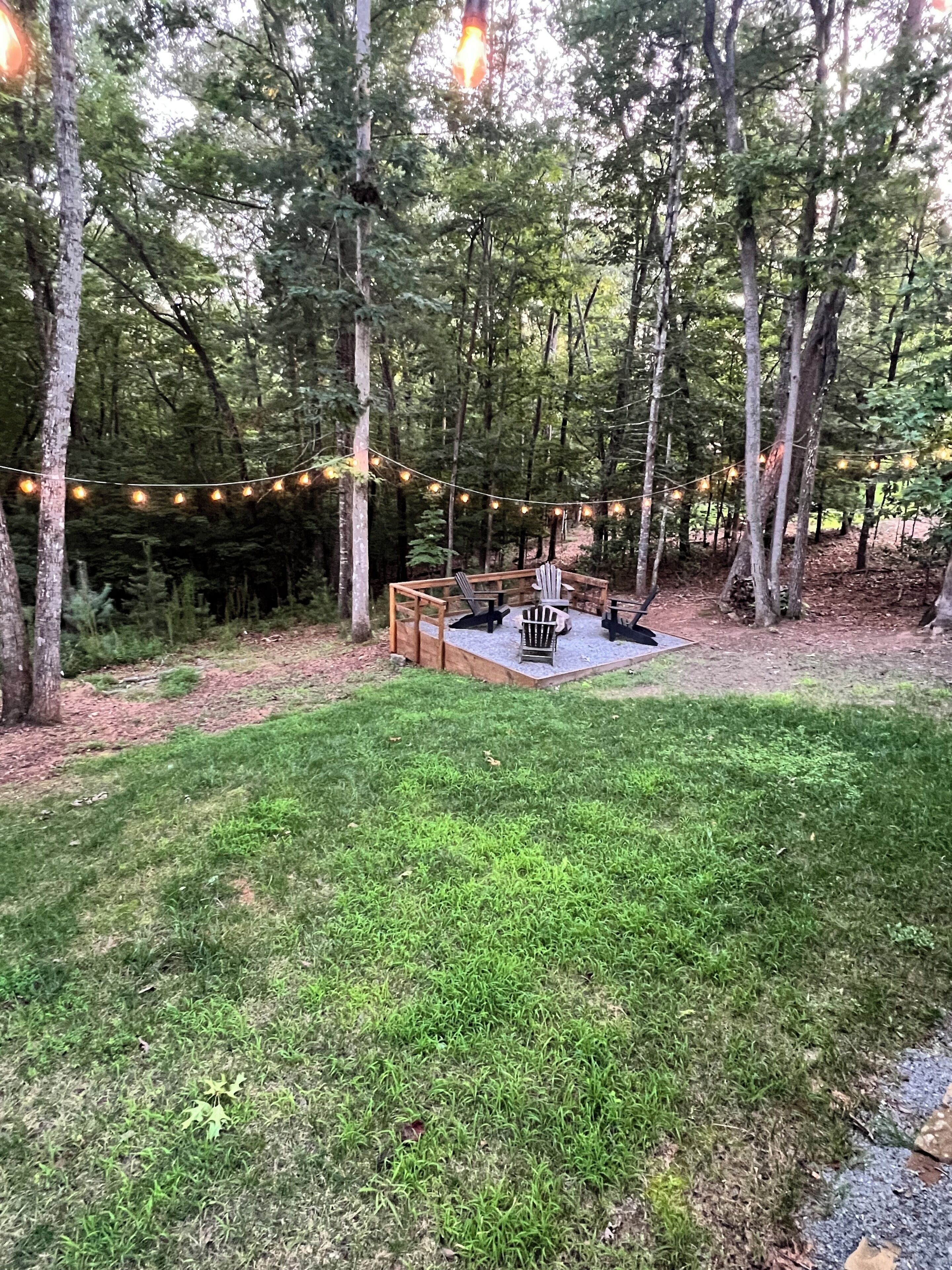 A forested outdoor area with a wooden deck, string lights, and outdoor furniture.