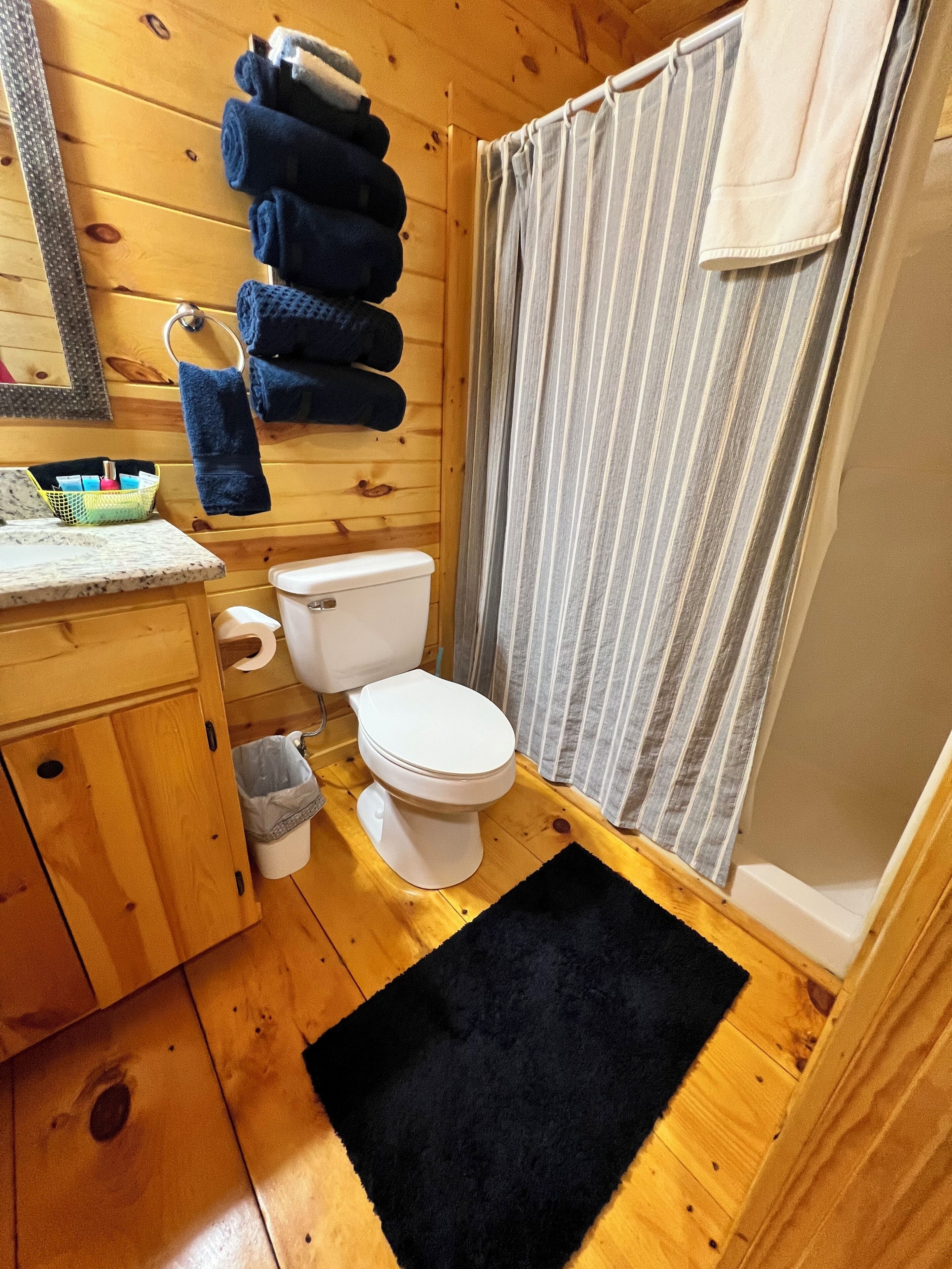 A compact wooden bathroom with a toilet, a shower curtain, a sink with a marble countertop, and a towel rack with folded towels.