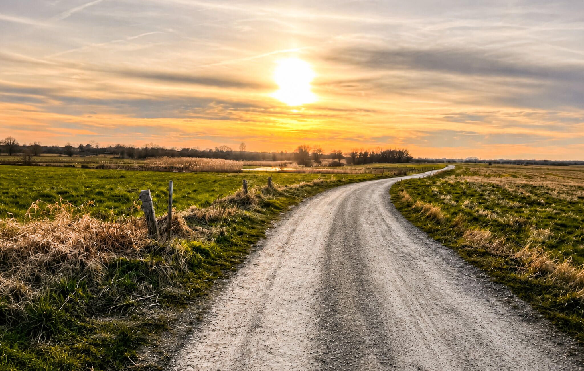 Un chemin de terre sinueux qui traverse des champs, avec un coucher de soleil en arrière-plan.