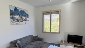A living room with a sofa, a coffee table, a TV, a painting, and a window with a view of greenery. - Le Canigou - FUILLA - Vacation Rental (FUILLA)