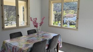 A dining area with a table covered by a leaf-patterned tablecloth, surrounded by chairs, and a vase with pink flowers. - Le Canigou - FUILLA - Vacation Rental (FUILLA)