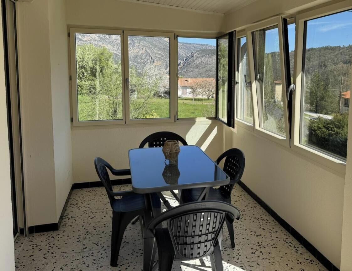A room with a table and chairs, large windows with a view of greenery and mountains, and a tiled floor.