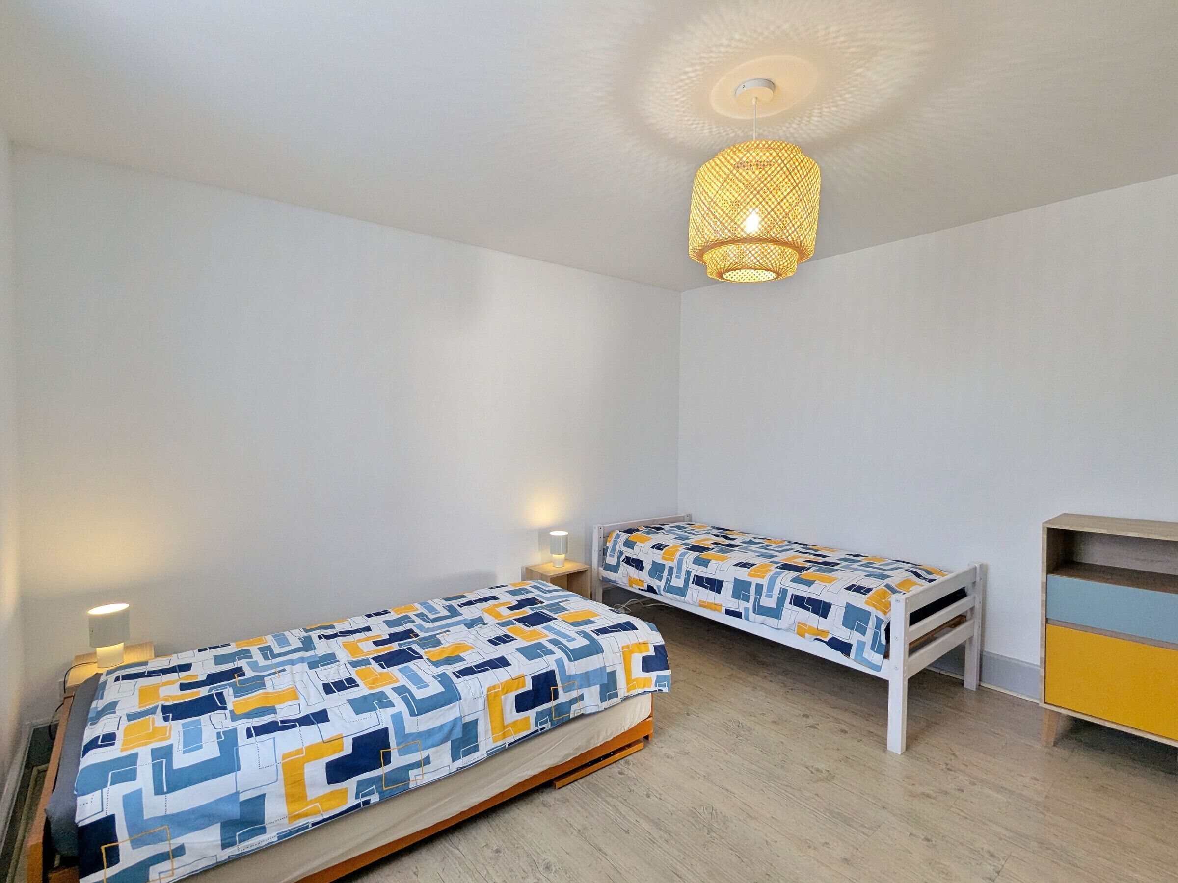 Un dormitorio con dos camas individuales, una mesita de noche con lámpara, un armario y una lámpara colgante.
