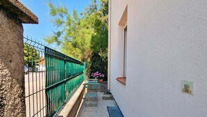 Une ruelle étroite, avec une clôture en métal vert, un mur blanc percé d’une fenêtre et un petit banc agrémenté de fleurs.