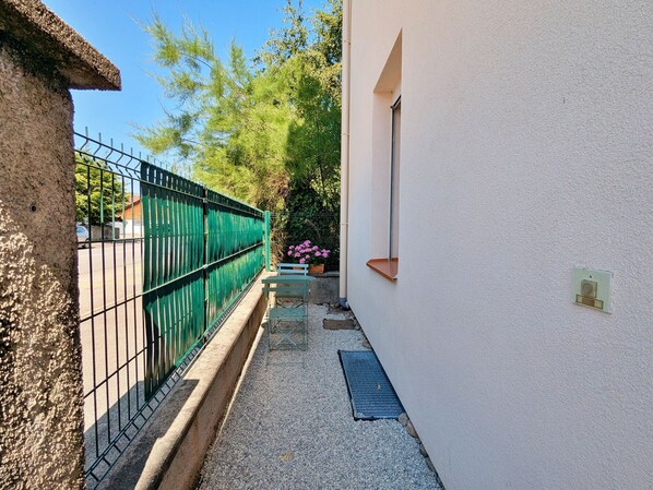 A narrow alleyway with a green metal fence, a white wall with a window, and a small bench with flowers.